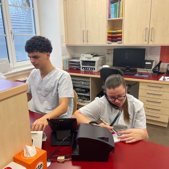 Zwei Personen in weißen Laborkittel arbeiten an einem Schreibtisch mit Monitor, Telefon und Drucker. Eine Frau benutzt ein Telefon und ein Mann schaut auf einen Computer.