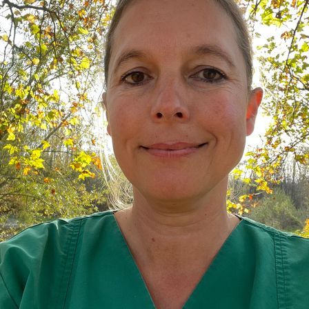 Eine Frau in einem grünen Scrub-Oberteil lächelt vor einem Baum mit gelben Blättern für die Kamera.