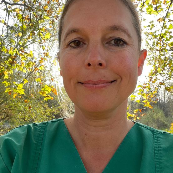 Eine Frau in einem grünen Scrub-Oberteil lächelt vor einem Baum mit gelben Blättern für die Kamera.