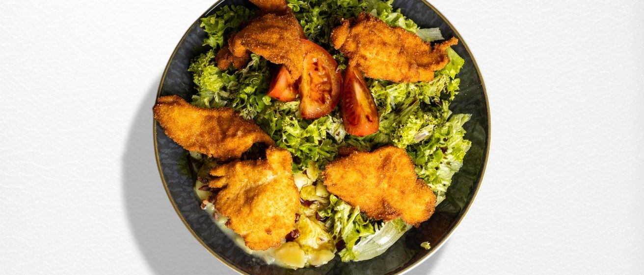 Eine Schüssel gemischter Blattsalat mit knusprigen gebratenen Hühnchenstücken und Tomatenscheiben auf weißem Hintergrund.