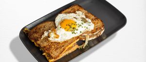 Zwei Scheiben getoastetes Brot mit geschmolzenem Käse und einem Spiegelei mit Zwiebeln und Schnittlauch auf einem schwarzen Teller.