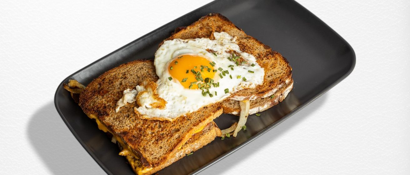 Zwei Scheiben getoastetes Brot mit geschmolzenem Käse und einem Spiegelei mit Zwiebeln und Schnittlauch auf einem schwarzen Teller.