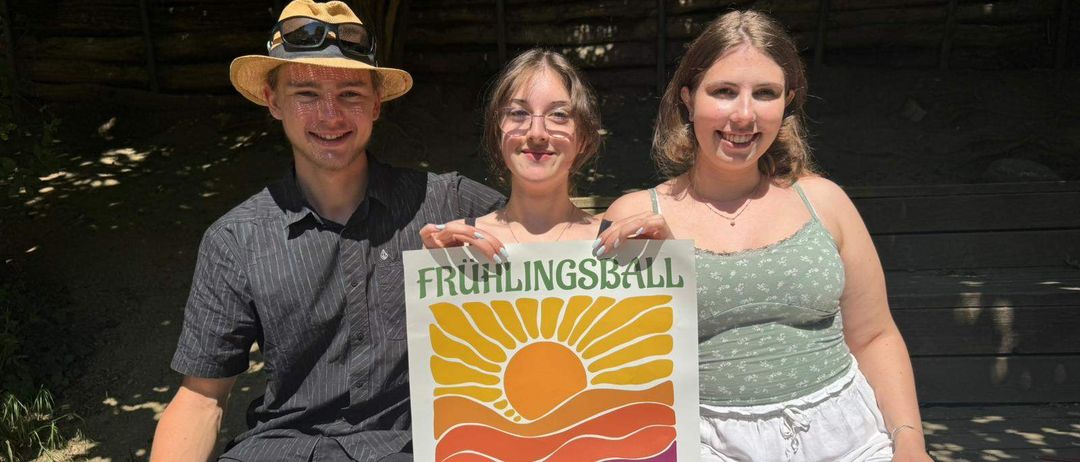 Drei Personen, zwei Frauen und ein Mann, sitzen auf einer Bank und halten ein Schild mit der Aufschrift Fruhlingsball Ball der 10. bis 12. Klasse der Waldorfschule Eisenstadt am 29. Mai 2026.