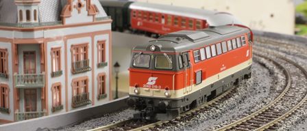 Ein roter und grauer Modellzug mit der Bezeichnung 2043 015-3 steht auf den Gleisen in der Nähe eines weißen Gebäudes mit mehreren Fenstern. Ein weiterer roter Zug ist dahinter.