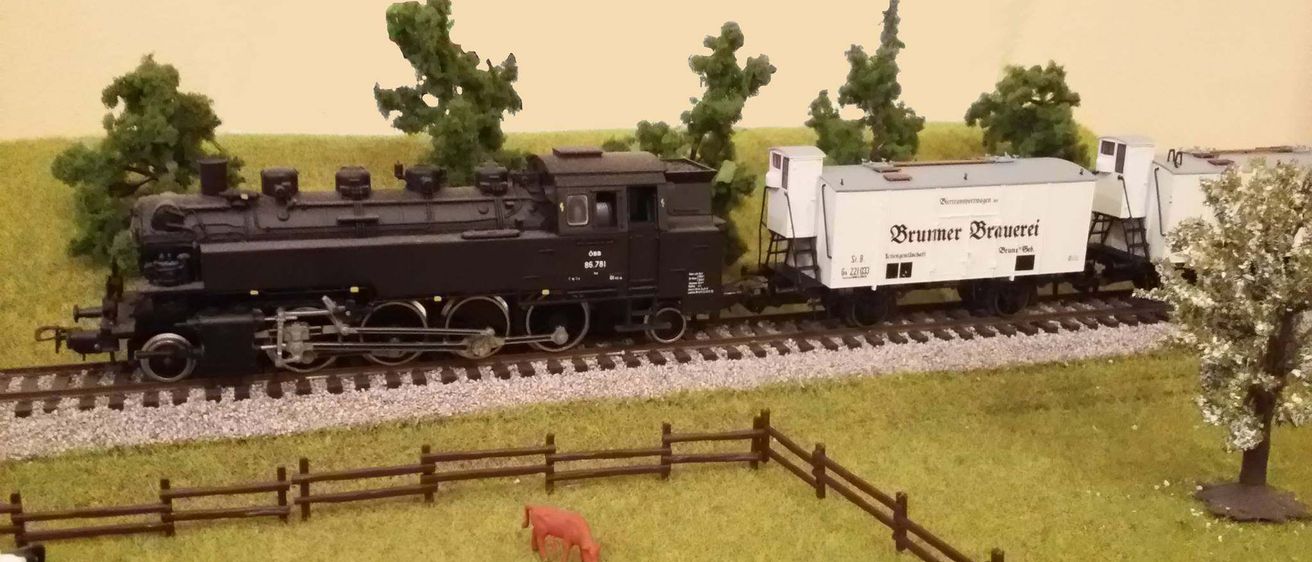 Ein Modellzug mit einer schwarzen Lokomotive mit der Aufschrift '86.781' und einem weißen Wagen mit der Aufschrift 'Brummer Brauer'. Zwei Kühe grasen im Gras in der Nähe eines Holzzauns.