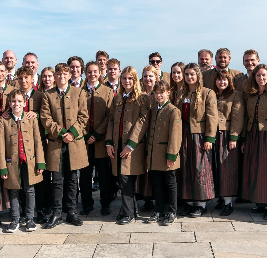 Eine Gruppe von Menschen in traditioneller bayerischer Kleidung, die auf einem Steinboden stehen, lächeln und für ein Foto posieren.