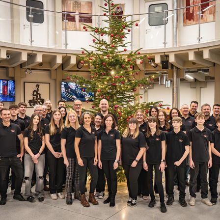 Eine große Gruppe von Mitarbeitern in schwarzen Hemden steht in einem modernen Büro rund um einen geschmückten Weihnachtsbaum, mit Monitoren und Kunstwerken im Hintergrund.