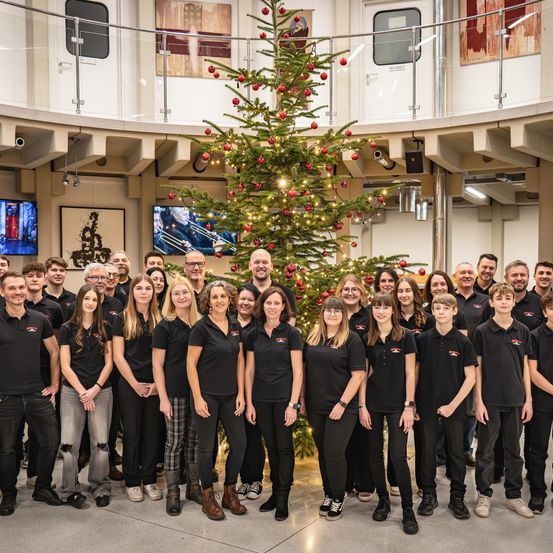 Eine große Gruppe von Mitarbeitern in schwarzen Hemden steht in einem modernen Büro rund um einen geschmückten Weihnachtsbaum, mit Monitoren und Kunstwerken im Hintergrund.