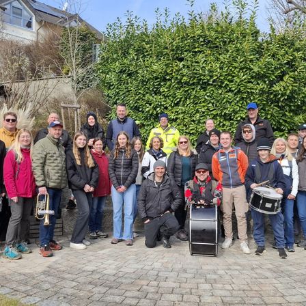 Eine Gruppe von Menschen steht im Kreis auf einem Ziegelboden. Einige halten Trommeln und Trompeten. Dahinter steht eine Hecke und ein Gebäude mit Solarpanels.