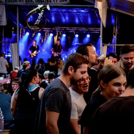 Ein nächtliches Konzert ist im Gange. Zwei Männer spielen Gitarren auf einer Bühne unter blauen Lichtern. Vor ihnen ist eine Menschenmenge versammelt, wobei ein Mann im Vordergrund einen Becher hält.