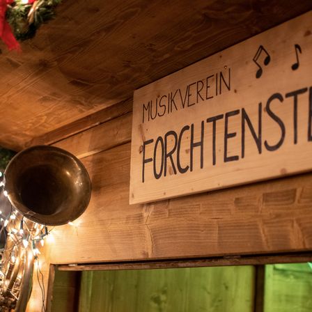 Ein Holzschild mit schwarzem Text 'Musikverein Forchtenstein' hängt an einer Holzwand, mit einem Messinginstrument und Weihnachtsdekorationen im Hintergrund.