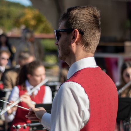 Ein Mann in einer roten Weste dirigiert eine Band, während er eine Sonnenbrille trägt und Taktstöcke in der Hand hält. Hinter ihm spielen mehrere Bandmitglieder ihre Instrumente, und der Hintergrund ist unscharf.