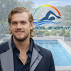 Ein lächelnder Mann steht vor einem Schwimmbecken und trägt eine graue Jacke und eine Halskette. Im Hintergrund befindet sich ein Logo mit einem Schwimmer und dem Text 'POOL-SHOP.AT'.