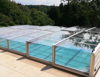 Ein Freischwimmbecken mit Glasdach und Metallrahmen, umgeben von Bäumen und Grün, mit klarem blauem Wasser.