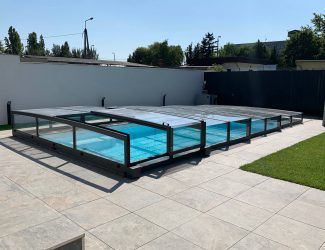 Ein Außenpool ist mit einem Glasdach bedeckt. Der Pool ist von einer Terrasse mit Rasenfläche und weißen Wänden umgeben. Im Hintergrund befinden sich Bäume und Gebäude.