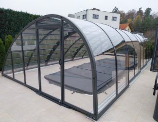 Ein modernes Glasgehäuse mit einem gebogenen Dach, auf einer Terrasse gelegen, verfügt über ein Bett im Inneren und ist von Pflanzen und einem Gebäude umgeben.