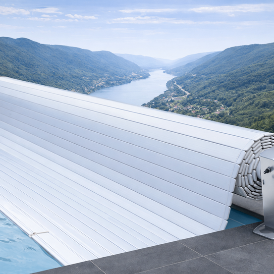 Eine weiße Poolabdeckung mit Spiraldesign steht am Rand eines Schwimmbeckens und bietet Blick auf einen ruhigen See und üppige Berge.
