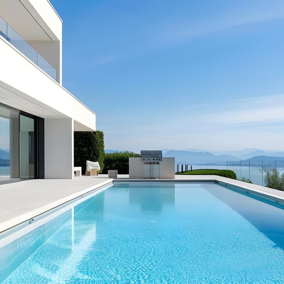 Eine moderne weiße Villa mit einem großen Pool, klarem blauem Wasser und einem Grillbereich. Der Pool ist von einer Terrasse umgeben, und im Hintergrund befinden sich Berge und ein See.