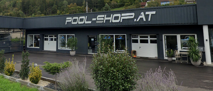 Die Außenansicht von POOL-SHOP.AT, einem Geschäft mit einem Berg im Hintergrund, zeigt Pflanzen, Sträucher und eine Einfahrt.