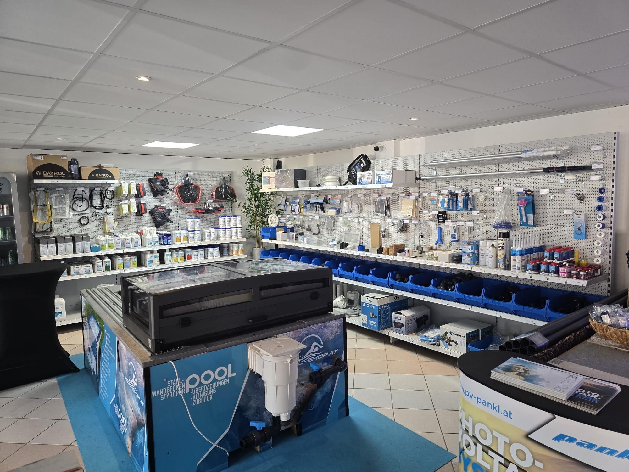 Ein Innengeschäft mit einem blauen Teppich, das eine Vielzahl von Produkten präsentiert, darunter ein großer schwarzer Aquarienheizer zur Schau gestellt ist, umgeben von Regalen, die mit verschiedenen Artikeln gefüllt sind. Die Decke ist weiß mit mehreren Lichtern.