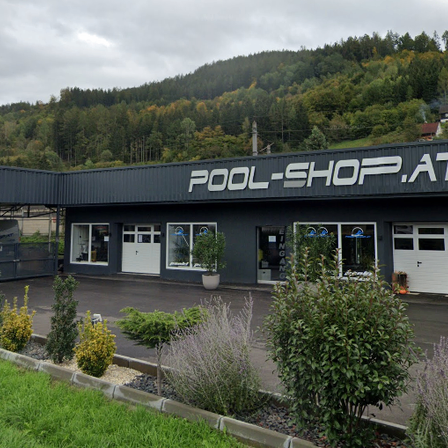 Das Pool-Shop.at-Gebäude mit grauer Außenfassade und weißen Texten befindet sich vor einem bewaldeten Hügel. Vor dem Gebäude stehen Pflanzen, Büsche und ein Rasen. Auf der linken Seite befinden sich ein Zaun und ein Tor. Der Himmel ist bewölkt.