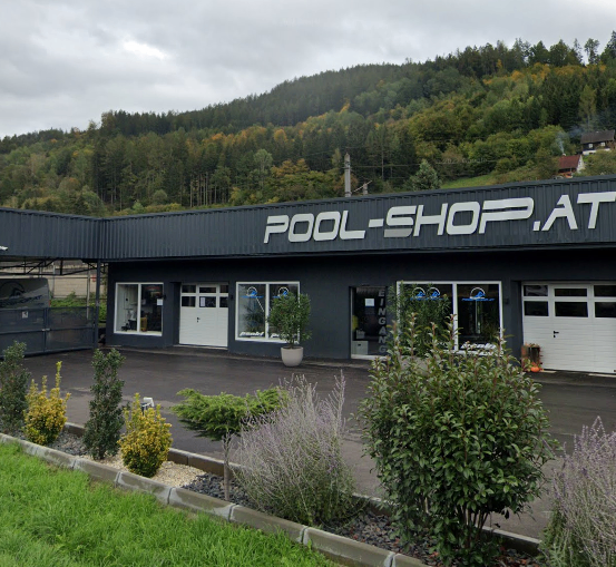 Das Pool-Shop.at-Gebäude mit grauer Außenfassade und weißen Texten befindet sich vor einem bewaldeten Hügel. Vor dem Gebäude stehen Pflanzen, Büsche und ein Rasen. Auf der linken Seite befinden sich ein Zaun und ein Tor. Der Himmel ist bewölkt.