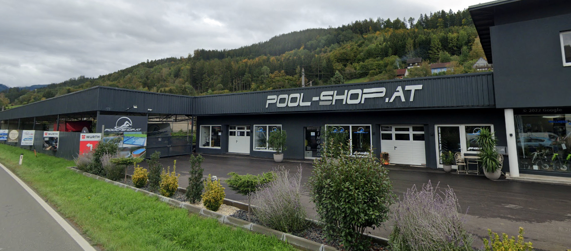 Das Pool-Shop.at-Gebäude mit grauer Außenfassade und weißen Texten befindet sich vor einem bewaldeten Hügel. Vor dem Gebäude stehen Pflanzen, Büsche und ein Rasen. Auf der linken Seite befinden sich ein Zaun und ein Tor. Der Himmel ist bewölkt.