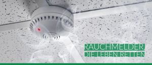 Ein Rauchmelder mit rotem Licht ist an einer weißen Decke mit weißem Gittermuster befestigt. Der Melder gibt Rauch ab und hat eine Website-Adresse, www.vers, in Grün.