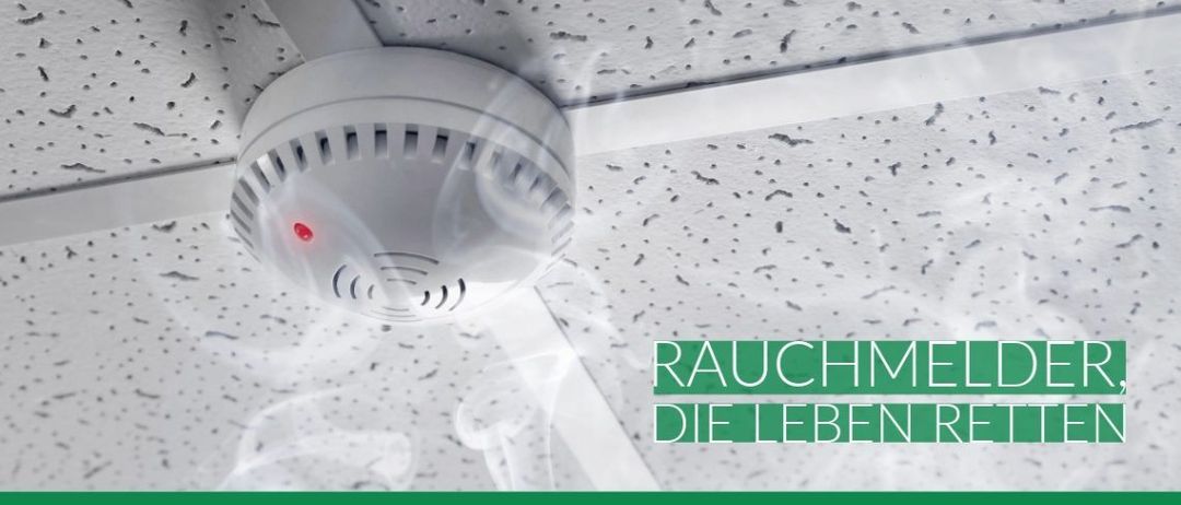 Ein Rauchmelder mit rotem Licht ist an einer weißen Decke mit weißem Gittermuster befestigt. Der Melder gibt Rauch ab und hat eine Website-Adresse, www.vers, in Grün.