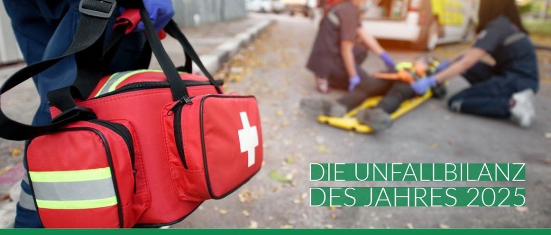 Sanitäter kümmern sich um einen Patienten bei einem Unfall, im Hintergrund ein Krankenwagen. Das Bild zeigt eine rote Sanitätstasche.
