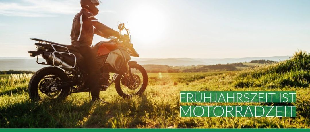 Ein Motorradfahrer steht auf einem Motorrad in einem Grasfeld mit Bergen im Hintergrund. Der Text lautet 'FRUHJAHRSZ MOTORRAI'. Unten befindet sich eine Website-URL.