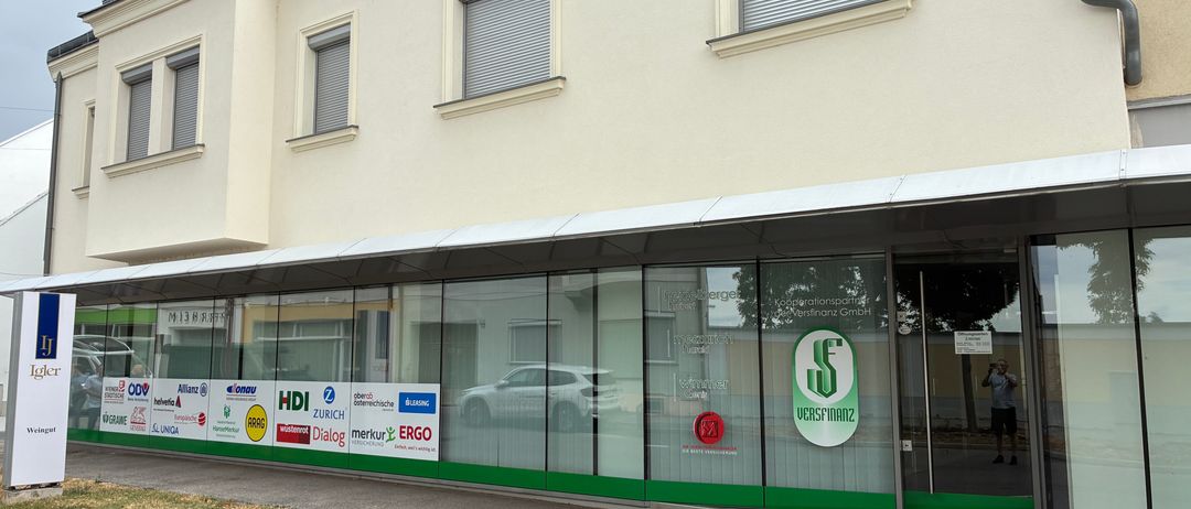 Ein Geschäftsgebäude mit einer Glasfassade und mehreren Logos an den Fenstern. Das Gebäude hat eine weiße Wand und ein graues Dach.