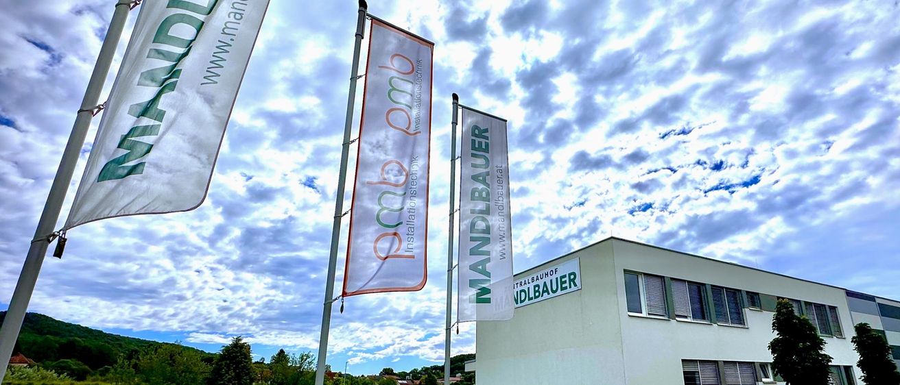 Ein weißes Gebäude mit einem Schild, auf dem 'Mandlbauer' steht, befindet sich in einem Rasenfeld unter einem bewölkten Himmel. Vor dem Gebäude stehen drei Banner. Ein grüner Lastwagen fährt auf der Straße vor dem Gebäude.