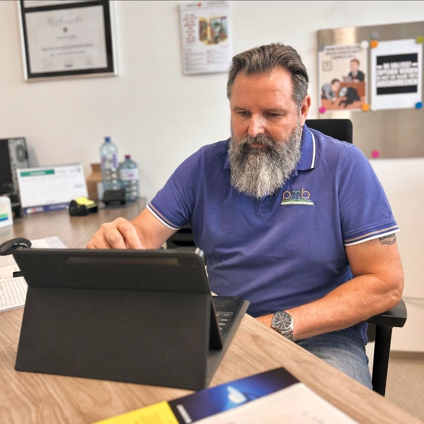 Ein bärtiger Mann in einem blauen Hemd sitzt an einem Schreibtisch und benutzt einen Laptop und ein Tablet. Papiere und ein Notizblock liegen auf dem Schreibtisch. Hinter ihm befindet sich eine Pinnwand mit Papieren und einem Zertifikat.