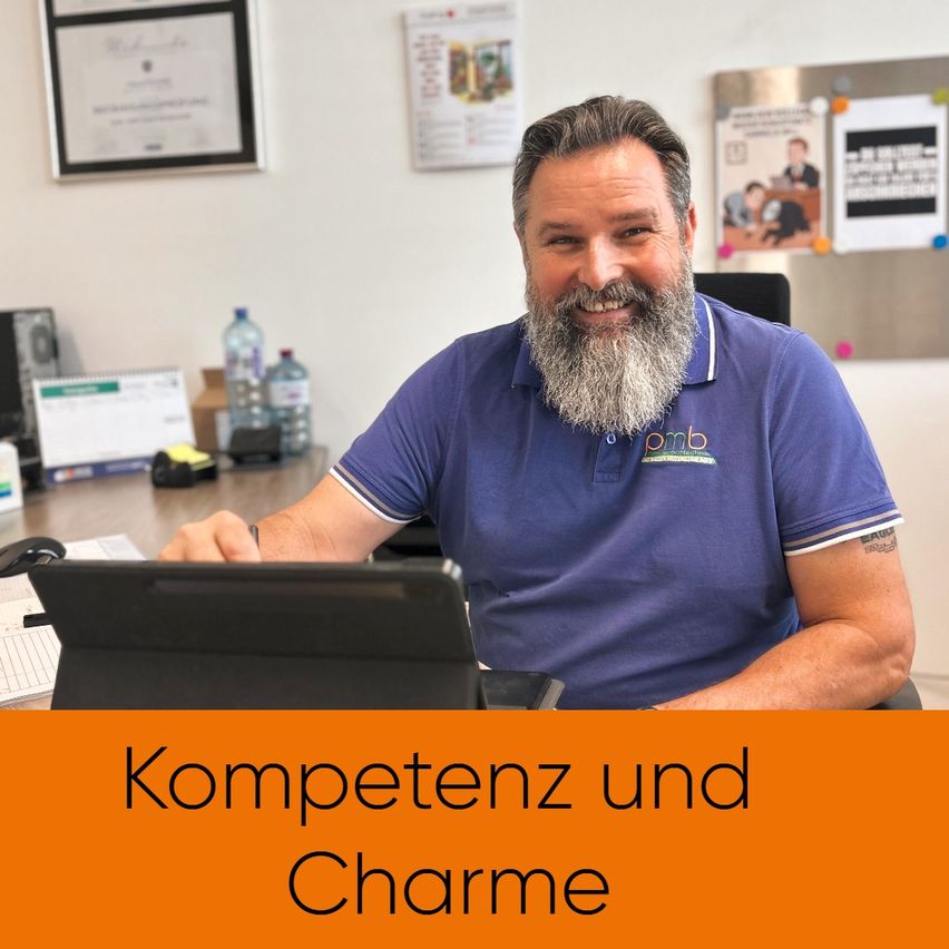 Ein lächelnder Mann mit Bart sitzt am Schreibtisch und trägt ein lila Poloshirt mit Logo. Hinter ihm befindet sich eine Pinnwand mit verschiedenen Papieren. Auf dem Schreibtisch stehen Flaschen, ein Notizblock und ein Computermonitor. Das Bild hat einen Textüberlagerung, der 'Kompetenz und Charme' lautet.