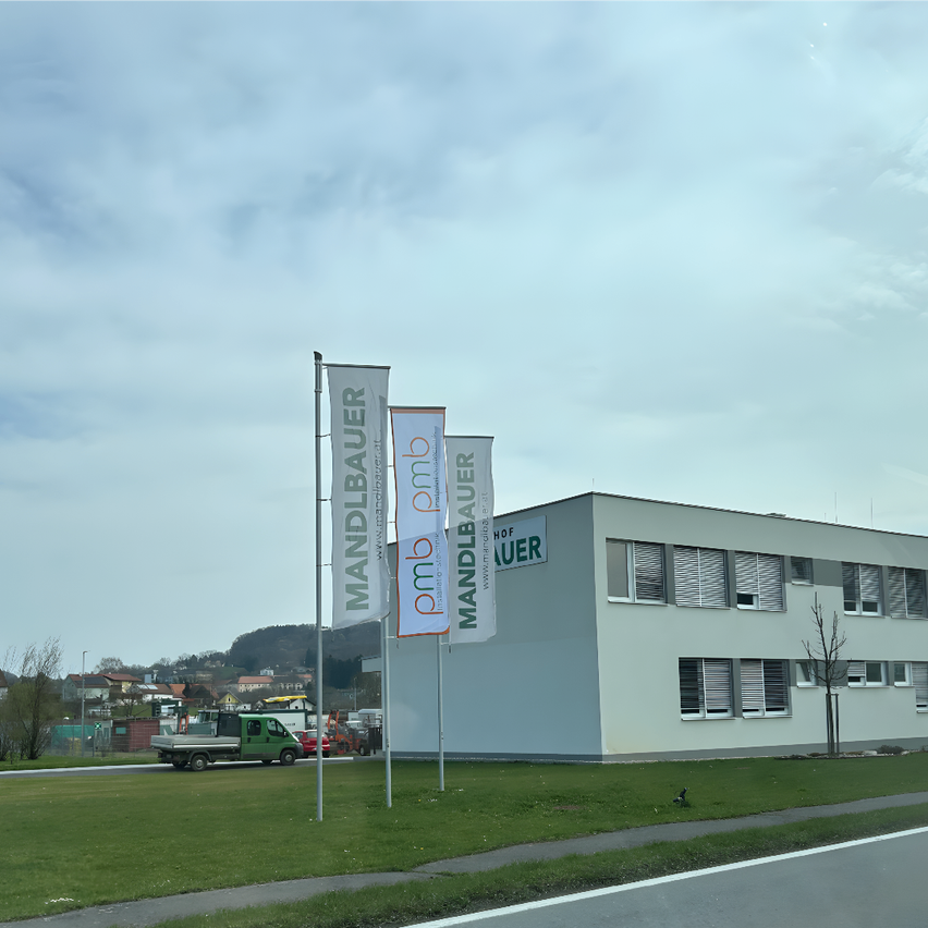 Ein Gebäude mit dem Namen Hof Mandelbauer ist zu sehen, mit mehreren Bannern davor. Ein grüner LKW ist vor dem Gebäude geparkt. Der Himmel ist bewölkt.