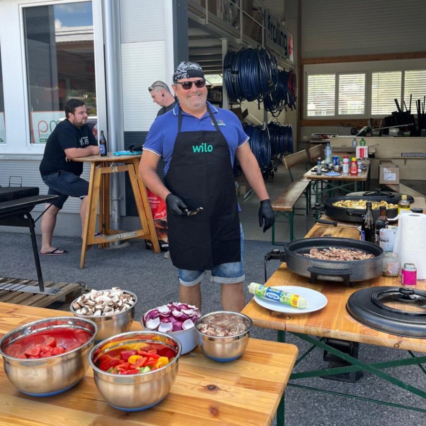 Ein Mann in einer Schürze steht an einem Tisch mit Schüsseln voller Gemüse und einem Grill. Hinter ihm sitzt ein anderer Mann auf einem Hocker, und es gibt mehr Gegenstände auf dem Tisch.