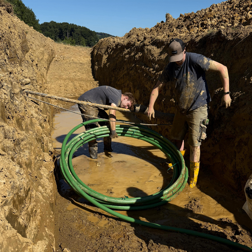 Zwei Männer arbeiten in einem schlammigen Bereich und handhaben eine große grüne Rohrleitung. Einer beugt sich vor, während der andere steht und das Rohr hält. Beide tragen Handschuhe und Stiefel.