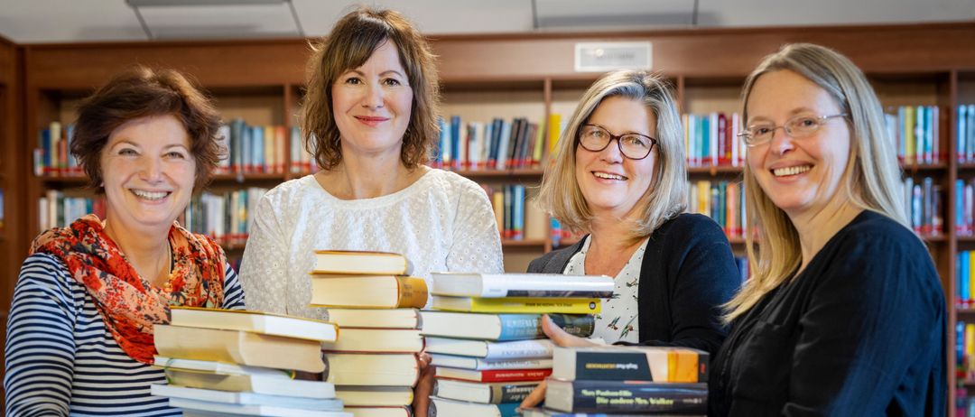 Vier Frauen lächeln und halten einen großen Stapel Bücher in einer Bibliothek.