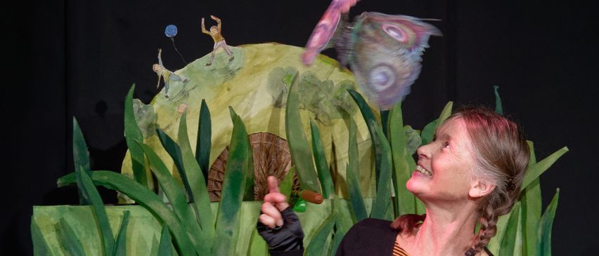 Eine Frau lächelt und zeigt auf eine große Schmetterlingspuppe, die über einer Bühne mit grünem Hintergrund und Figuren fliegt.