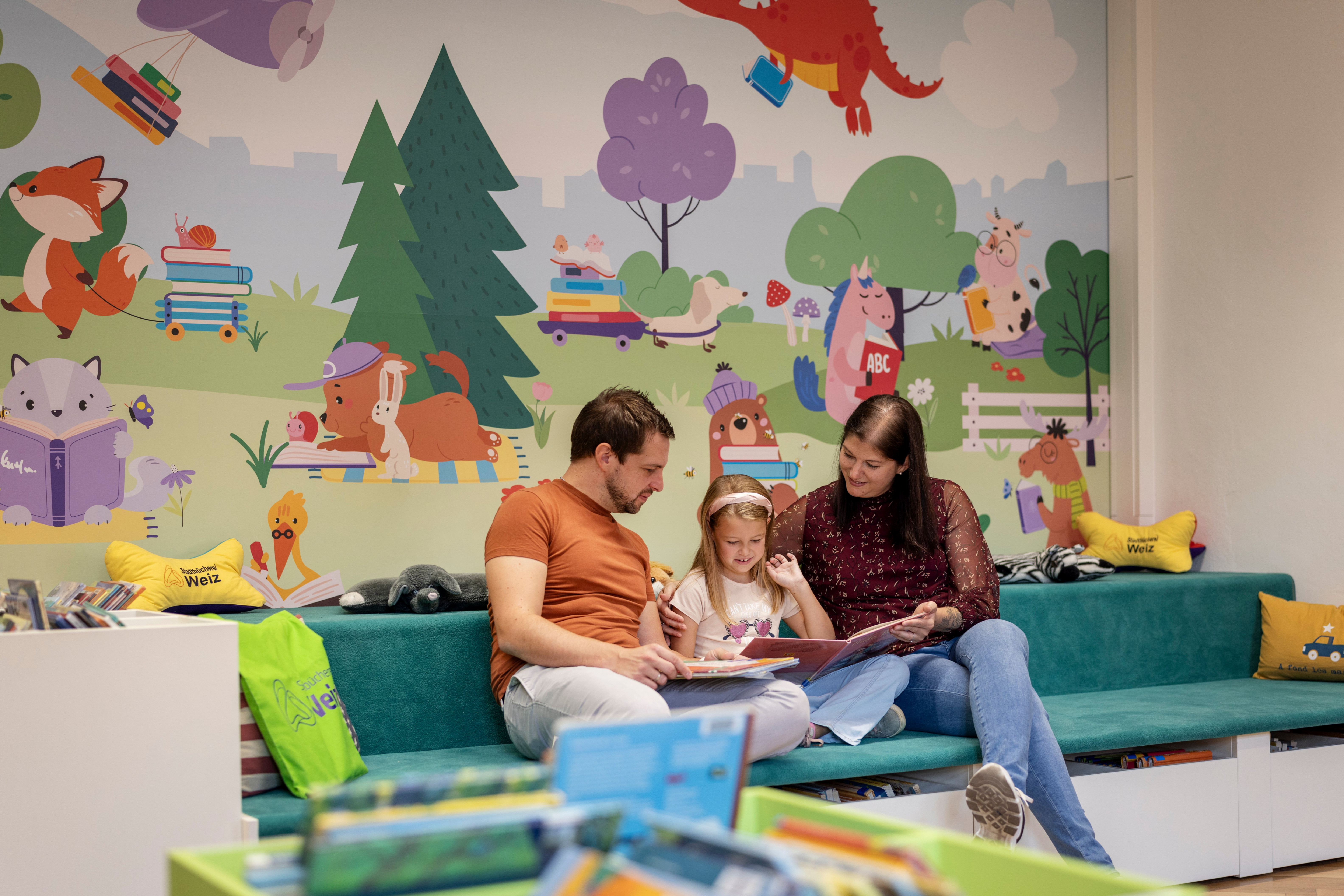 Familie liest in der Stadtbibliothek Weiz ein Buch vor einer bunten Wandmalerei.