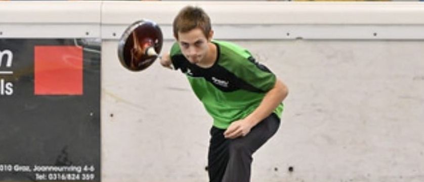 Ein junger Mann in einem grünen Shirt wirft einen Frisbee. Zwei Personen beobachten ihn hinter einer Barriere.