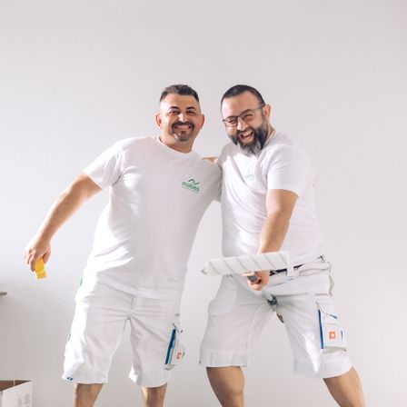 Zwei Männer in weißen Uniformen und mit Brille stehen vor einer weißen Wand, lächelnd halten sie einen Farbroller und eine Farbdose.