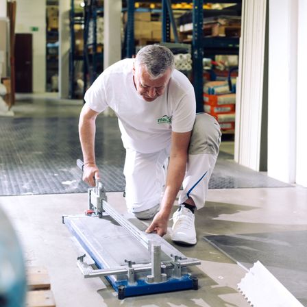 Ein Mann in einem weißen T-Shirt und Hose arbeitet in einem Lager und beugt sich vor, um einen Fliesenschneider zu benutzen.