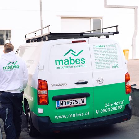 Ein weißer und grüner Van mit dem Text mabes und einem Logo steht in einer Garage. Ein Mann in einem weißen Hoodie steht dahinter.