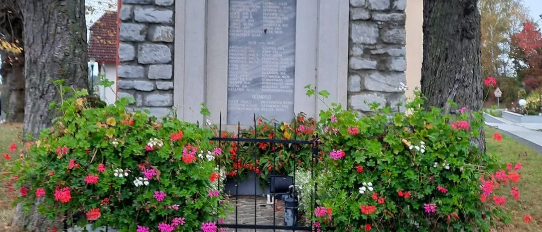 Ein kleines Steindenkmal mit einem roten Dach und einem Kreuz darauf. Es ist von einem schwarzen Zaun und Blumen umgeben.