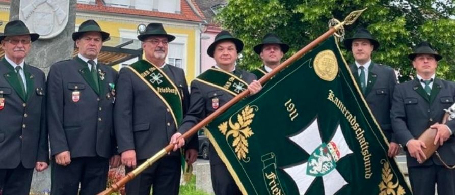Sieben Männer in grünen Uniformen und Hüten stehen vor einem Denkmal. Einer hält eine Fahne mit einem Emblem, und die anderen tragen Musikinstrumente. Ein großer Baum und Gebäude stehen hinter ihnen.