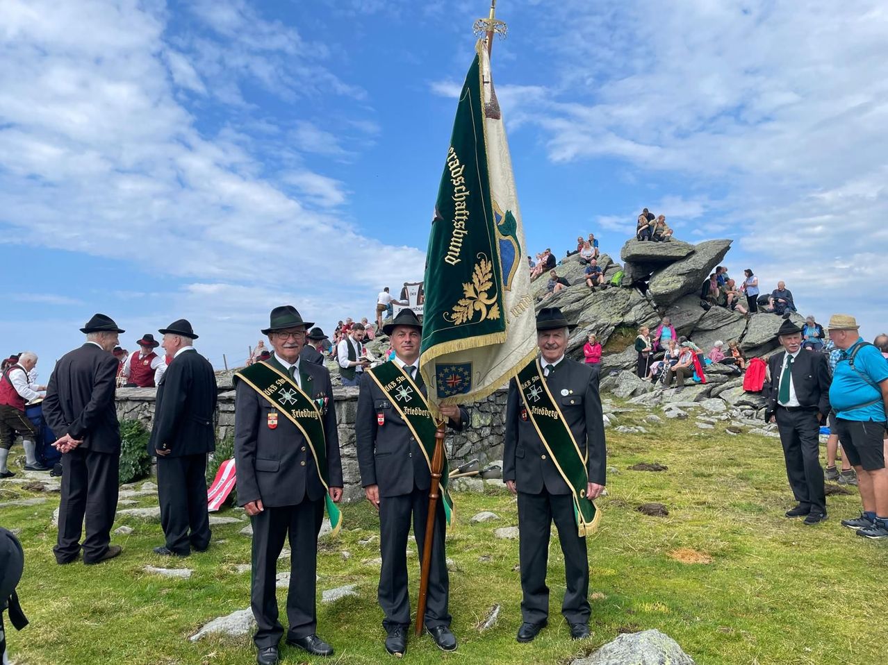 Drei Männer in Uniform, einer hält eine Fahne, stehen auf einem Grashügel mit einer Menschenmenge im Hintergrund.