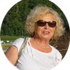 Eine ältere Frau mit blonden, gelockten Haaren, Sonnenbrille und einem weißen T-Shirt steht draußen. Im Hintergrund ist ein Wald zu sehen.