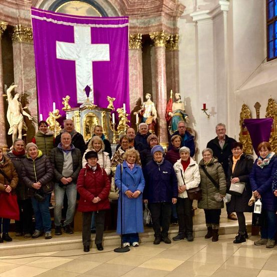 Eine Gruppe von Menschen steht vor einem Altar in einer Kirche, mit einem violetten Banner mit einem Kreuz im Hintergrund.